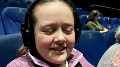 Eryn has long brown hair and is pictured at the cinema in a pink fleece and is wearing an audio descriptive headset. The cinema seats are all blue and a few people are seen in the cinema in the background. 