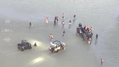 Two cars in the shallow water with people surrounding what appears to be a dingy boat with people inside the vessel