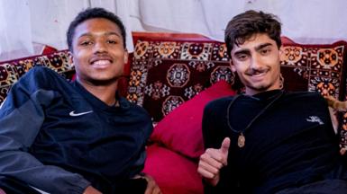 Jo and Kush smile as they lean back on maroon patterned cushions in a Turkish home.
