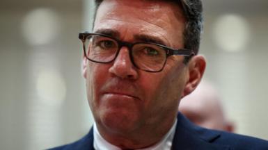 Andy Burnham looks directly forward, his lips are purse and he wears tortoise shell glasses. His suit jacket is navy and his shirt is white. The background is blurred grey.