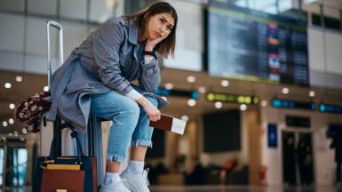 Young woman wearing blue jeans, a grey trenchcoat and white trainers sits on her suitcase at an airport, clutching her passport and ticket 