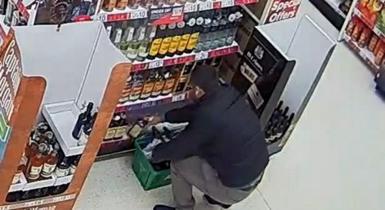 CCTV image of a man in a black jacket in jeans, crouching down on a supermarket floor, next to shelves of alcohol bottles. He is putting them into a green shopping basket.