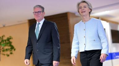 A man in a suit and blue tie strides forward beside a woman in dark trousers and a light blue jacket