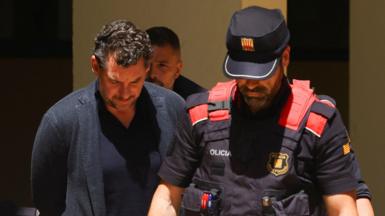Jonathan Andic wears a blue jacket and blue top while he walks with his hands behind his back and his head down. He stands next to a policeman who is wearing a black and red uniform and cap.