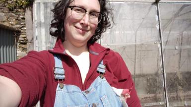 Alix Harvey, wearing glasses and a red fleece with denim dungarees, smiles in a selfie 