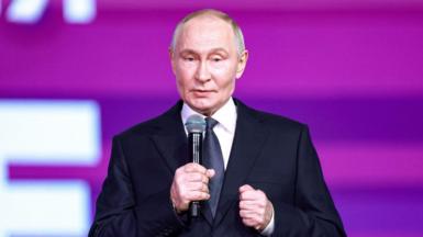 Putin, wearing a suit and tie, speaks into a microphone in front of a purple board.