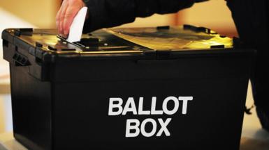 A voter placing a ballot paper in a black ballot box at a polling station.