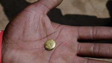 A tiny gold ingot sits on the on the palm of a hand in close-up. A wrist band saying "World Aids day" is also visible.