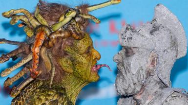 Woman dressed as a gorgon from Greek mythology, with snakes for hair and coloured green, sticks her tongue out at a man, who is grey and appears to have turned to stone.