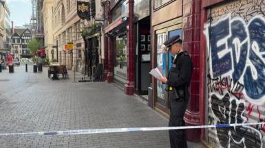 A police officer stands behind police tape on a city street in central London, reading notes outside a closed shop with graffiti on its shutter, while nearby streets are quiet and cordoned off.