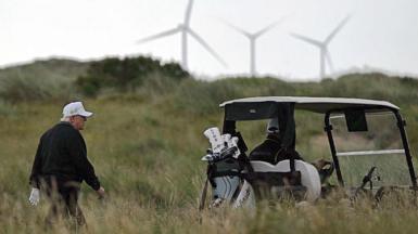 President Trump in a white cap walks towards a golf buggy with wind turbines in the distance