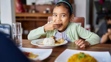Cute girl looking away while eating food at home. Female is having lunch at dining table. She is in casuals.
