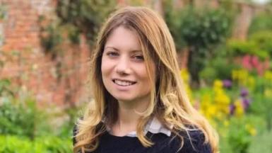 Rebecca Ableman smiling to the camera in a dark navy knitted jumper - there is a beautiful walled garden behind her