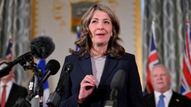 An image of Alberta Premier Danielle Smith standing and speaking at a podium. She is wearing a dark navy blue blazer with a tan top underneath and a silver necklace. She has shoulder-length brown, wavy hair. 