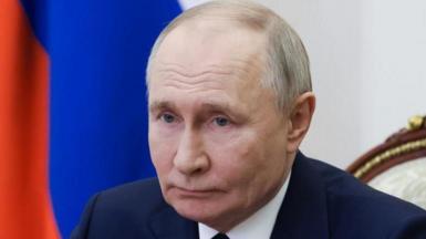 Head and shoulders picture of Vladimir Putin, wearing a dark suit and matching tie, with a Russian flag behind him.