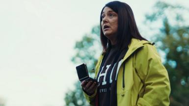 A woman confronts Mark Daly in the park, holding a mobile phone in her right hand. She has long brown hair and is wearing a yellow jacket and a dark grey jumper.