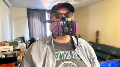 Brennah Hammar wearing the mask that she sleeps in and sunglasses, standing inside her apartment. There are stacks of items on a dresser, and bare yellow walls