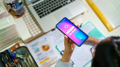 A young female student seen from above interacts with an AI chatbot on a smartphone while studying at a desk with a laptop, notes and stationery. The scene highlights modern learning and technology integration.