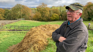 Farmer Alan French, wearing a cap and brown jacket, looks out at his field with his arms folded. He is standing by a fence and some hay.