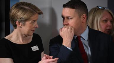 Amanda Pritchard, wearing a black top with a name tag on, stands speaking to West Streeting, who is wearing a suit. Pritchard is gesturing with her hands and Streeting is covering his mouth with his hand