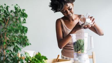 Stock photo shows a woman adding chia seends into a smoothie at home, there is a plant in the background.