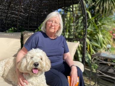 Avril is sitting outside in a blue top. She is sitting on a black wicker chair. Beside her is Dougie who is a white dog. Avril is facing the camera. Over the image is Your Voice, Your BBC New