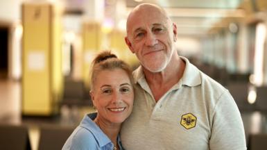 Two adults hugging side by side, one is a women wearing a sky blue shirt and the other ois a man wearing a beige t-shirt with a collar. Both are smiling at the camera