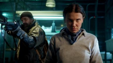 (L to R) David Harbour as Jim Hopper holding a gun and Millie Bobby Brown as Eleven looking straight at camera in Stranger Things.