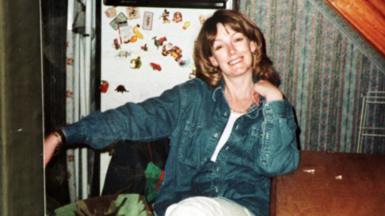Arlene Fraser sitting with her left elbow resting on a wooden table. She is wearing a blue denim shirt, white trousers and a white top. Behind her can be seen a fridge with a number of fridge magnets, patterned wallpaper and a wooden beam.
