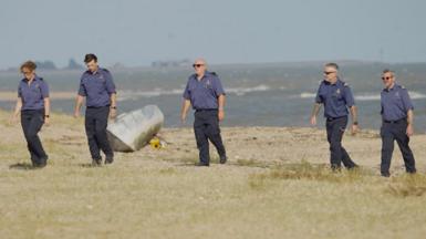 Five people wearing navy shirts and trousers walking in a line along the sand. The sea is behind them and there is a bouquet of yellow sunflowers on the sand.