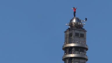 Alex Honnold stands on top of the Taipei 101 skyscraper building in a red t-shirt and dark trousers. There is only blue sky behind him. 