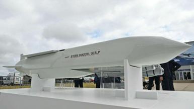 A Storm shadow missile stands against the backdrop of some grey clouds with people wandering around looking at it from the opposite side of the camera.,