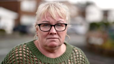 Mandy - a woman with blond hair, gold earrings and black-rimmed glasses. She is wearing a dark-green sweater. She is standing on a street.