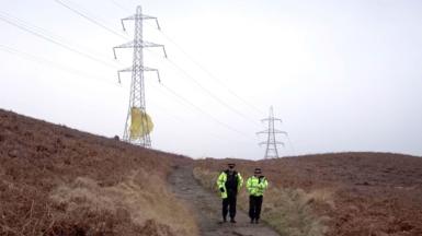A male and a female police officer walk down a remote dirt track, which is flanked on both side by gorse. On the horizon are two large electricity pylons, there is yellow-coloured material that is wrapped around the bottom of the nearest pylon that appears to be billowing in the wind 