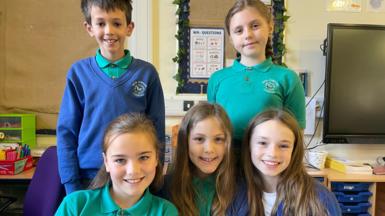 Five pupils wearing green polo shirts in two rows in a classroom with two pupils standing at the back and three girls sitting in front of them. There is one boy standing, wearing a  blue jumper. All the children are smiling. 