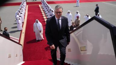 Keir Starmer boarding a plane at the airport in Doha, Qatar. There is a red carpet, lined with men, leading up to the plane