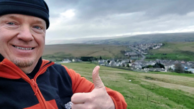 Joe Albon looks towards the camera in the middle of a field, with green countryside behind him. He is wearing a black hat, and a black and orange jumper. He is holding his thumb up and smiling at the camera.