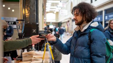 A male customer in a winter coat pays using contactless card payment at a station coffee stall, with the attendee taking payment out of shot save for an arm and a hand holding the card reader.