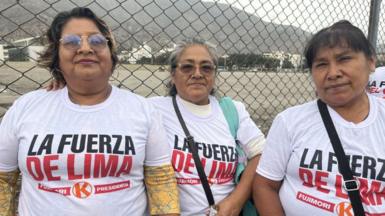 Three women wearing white Fujimori T-shirts, standing in front of a fence with mountains visible behind