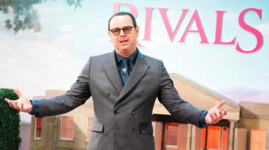 Danny Dyer in a grey suit holding his arms out in front of Rivals branding at the premiere