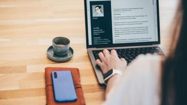 Back shot of a young woman working on her CV on a lap top on a wooden table with a cup of coffee, her mobile phone and a brown leather notebook next to her.