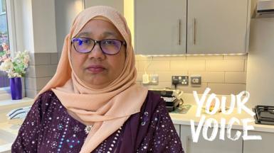 A woman in a peach hijab and glasses stands in a modern grey-hued kitchen. 