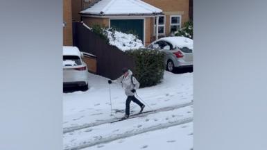 A man skiing down a residential street. He is wearing a grey jacket and black trousers.