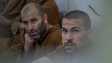 Two inmates sit on the ground against a wall both dressed in brown clothes