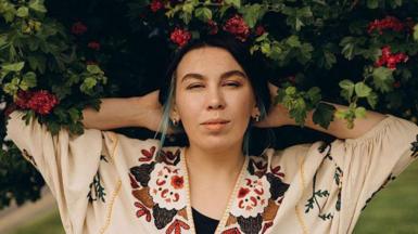 Nastya Zukhvala next to a leafy bush with red flower looking directly into the camera. 