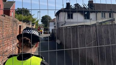 A police officer is stood facing a badly burnt property. The roof has caved in and exposed the wooden beams. An alley runs down next to the property and it has a wooden fence on one side