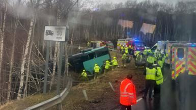A green bus at an angle down an embankment with lots of emergency service workers wearing high vis vests, ambulances and fire engines visible on the right hand side of the photo 