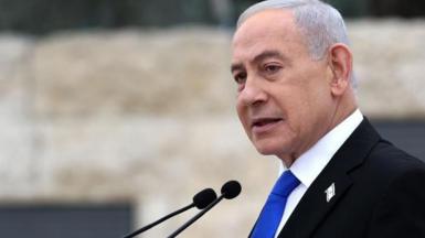 Netanyahu, who has grey short hair and wears a black suit with a blue tie and has a badge of the Israeli flag on his lapel, speaks into a mic at a podium