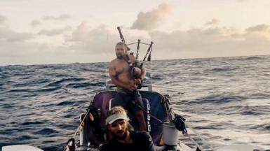 A man in bare chest on a small boat in the sea plays the bagpipes.