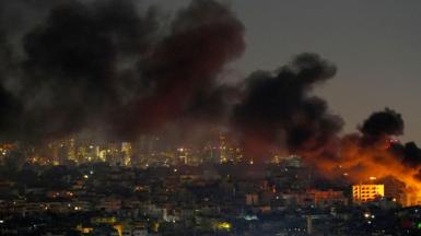 Smoke rises across the skyline of southern Beirut, appearing to come from a building glowing orange on the far right of the scene.